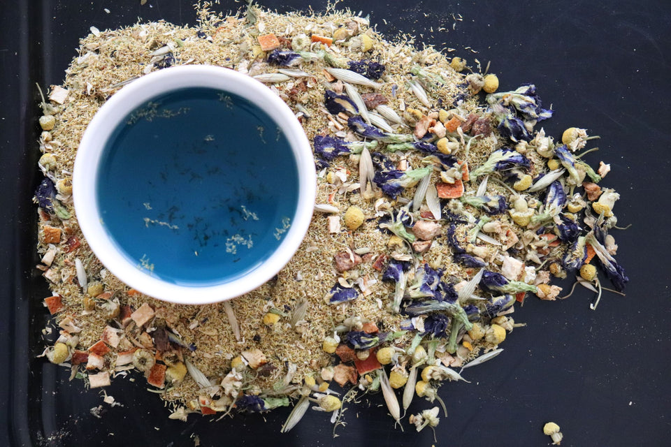 Herbal tea tinted blue from dried blue butterfly pea flowers, surrounded by dried orange peel, chamomile, and milky oat tops.
